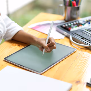 Person using a stylus on a tablet at a well-lit desk with art supplies – A close-up of someone sketching or designing digitally, surrounded by creative tools like colored pencils and notebooks.