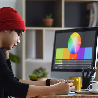 Young designer in a red beanie working at a computer with a colorful color wheel on screen and sketching notes beside it.