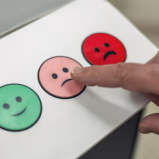Close-up of a person pressing a pink sad face button on a feedback panel, with green happy and red angry faces visible beside it.