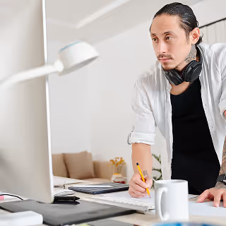 Focused man with headphones around his neck standing at a desk, writing with a pencil while looking at a computer monitor in a bright workspace.