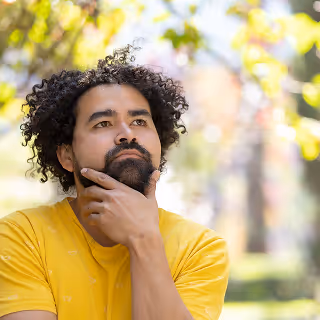 Man with curly hair and a beard wearing a yellow shirt, sitting outdoors thoughtfully with his hand on his chin, surrounded by blurred greenery.