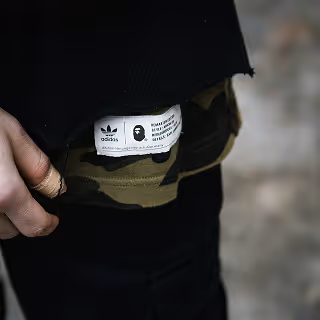 Adidas x BAPE camouflage detail – Close-up of a person’s hip wearing a camouflage Adidas x BAPE garment, showing the collaboration label tag under a black shirt.