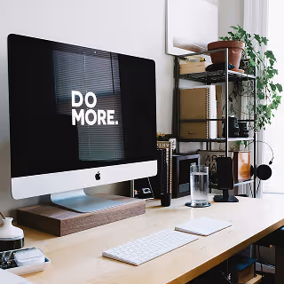 A desktop workspace featuring an iMac with the words “DO MORE.” displayed on the screen, surrounded by neatly arranged office supplies, books, and a small indoor plant.