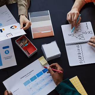 Overhead view of a group of people working together at a table with notebooks, charts, color swatches, and documents, collaborating on a creative project.