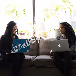 Two women sitting on a couch with laptops, smiling and engaging in a conversation, with bright natural light and potted plants in the background.