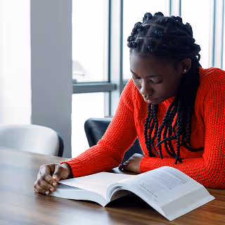 A woman in a red sweater with braided hair is deeply focused on reading a textbook at a wooden table near large windows, indicating a quiet study environment.