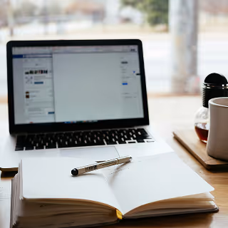 An open notebook with a pen placed on it sits in front of a laptop on a wooden desk, with a coffee mug and carafe nearby, suggesting a workspace for writing or content creation.