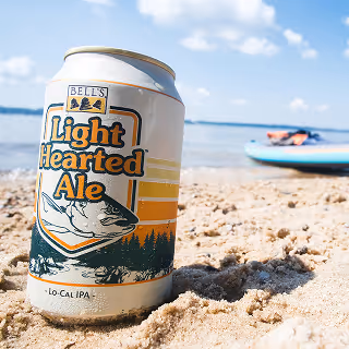 A can of Bell’s Light Hearted Ale resting on a sandy beach near water with a paddleboard in the background, capturing a relaxed summer vibe.