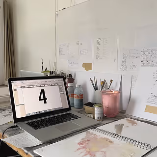 A minimalist creative workspace featuring an open laptop showing a large number "4" on the screen, surrounded by sketchbooks, stationery, and a lit candle on a white desk with concept notes taped to the wall.
