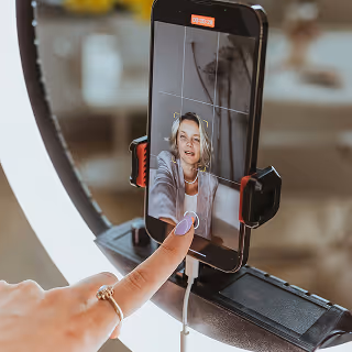 Close-up of a woman recording a selfie video using a smartphone mounted in a ring light setup, with her face visible on the phone screen and a finger about to tap the record button.