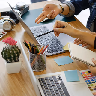 Close-up of hands working at a shared desk with laptops, sticky notes, and color swatches, suggesting collaborative creative work.