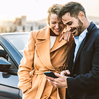 Smiling man and woman standing next to a car, warmly dressed, looking at a smartphone together, possibly checking information or reviewing something online.