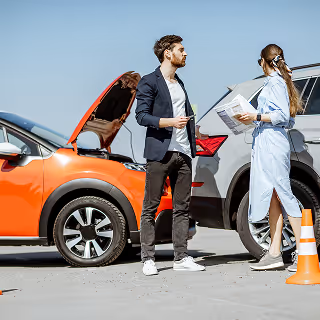 Two people standing between two cars that appear to have been in a minor accident; the orange car has its hood open, and traffic cones are placed around the scene.