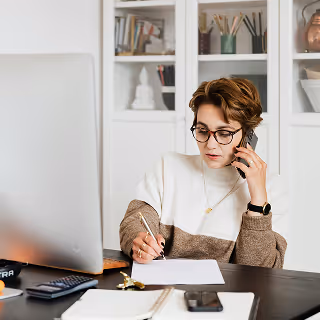 Woman in glasses multitasking at a desk, speaking on the phone while writing notes, with a computer and open notebook in front of her.
