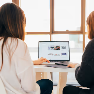 Two women sit side by side at a white table, reviewing an ecommerce website on a laptop in a sunlit modern office.