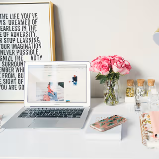 Minimalist desk setup with an open laptop displaying a fashion website, surrounded by pink roses, framed quote, and a smartphone.