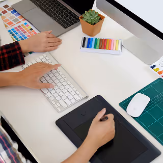 Overhead view of a designer using a keyboard and drawing tablet at a clean desk, surrounded by color swatches, an iMac, and pastel markers.
