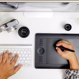 Top-down view of a designer's desk with a graphics tablet, keyboard, and camera lenses, showing hands actively working on a digital project.