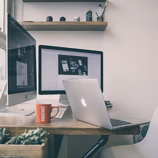 Minimalist desk setup with an Apple laptop and external monitor, coffee mug, and window light, suggesting a professional design environment.