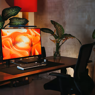 Stylish home office setup with a large monitor displaying abstract orange artwork, surrounded by plants and warm lighting.