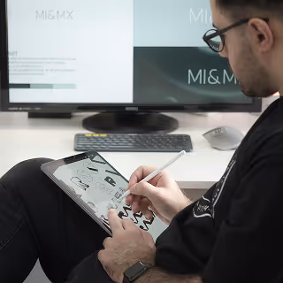 Man using a stylus on a tablet while sitting in front of a large monitor showing branding work, indicating digital design or logo creation.