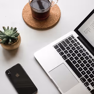 Overhead view of a workspace with a laptop, a smartphone, a potted succulent, and a glass mug of black coffee on a coaster.
