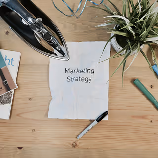 A crumpled sheet of paper labeled "Marketing Strategy" is placed on a wooden desk alongside a pen, iron, notebook, and a potted plant, suggesting creative planning or brainstorming.
