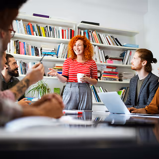 A woman with curly red hair stands smiling and holding a coffee cup while leading a discussion with four colleagues around a conference table in a modern office with bookshelves in the background.