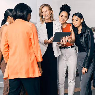 A group of diverse women in business attire gathered around a digital tablet, smiling and discussing ideas in a modern office space.
