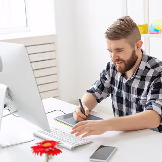 A man with a beard and checkered shirt working on a desktop computer using a pen tablet, smiling in a bright, minimal office.