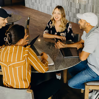 Diverse group of young professionals gathered around a round table, engaged in a lively discussion with laptops open in a modern workspace.