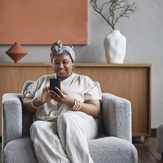 Woman relaxing on a gray armchair while smiling at her smartphone, with modern decor including a vase and abstract artwork in the background.