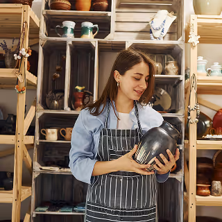 Smiling woman in striped apron admiring a handcrafted ceramic vase in a cozy, rustic pottery store filled with shelves of artisan wares.