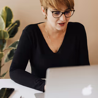 Woman with glasses and short hair wearing a black top, focused on her laptop during a virtual meeting.