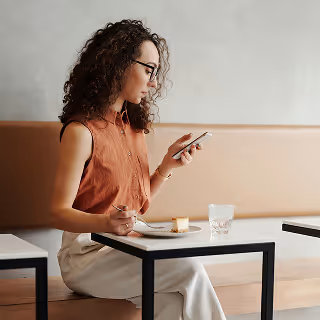 Woman with curly hair seated at a cafe table, eating dessert and reading something on her smartphone.