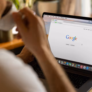 Person holding a cup and browsing Google homepage on a laptop in a cozy indoor environment.