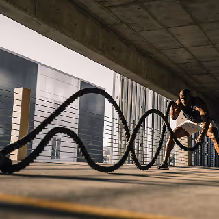Athletic man performing an intense workout with battle ropes under a covered outdoor area, with urban architecture in the background.