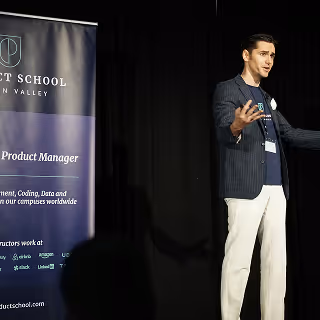 A speaker gesturing energetically on stage beside a "Product School" banner.