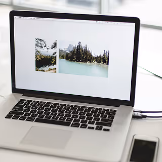 Open laptop on a white desk displaying landscape photos of lakes and pine forests, with a smartphone placed beside it.