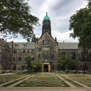 Grand stone university building with a tall central tower and green-topped spire, surrounded by neatly manicured lawns and trees.