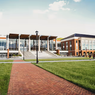 Contemporary university campus building with large glass windows, a central staircase, and a well-kept brick pathway leading through a grassy quad.