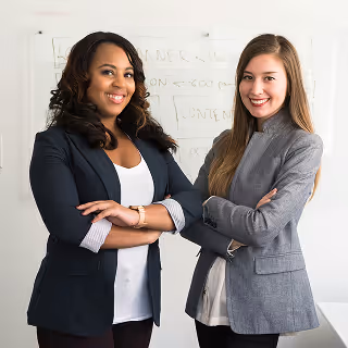Two women in professional blazers stand side-by-side with arms crossed, smiling in front of a whiteboard filled with marketing terms.