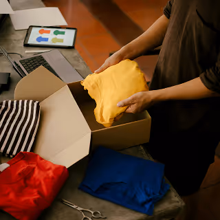 Person packing colorful t-shirts into a cardboard box on a table with a tablet, laptop, and scissors.