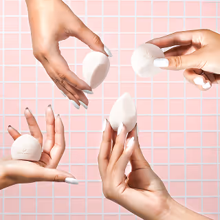 Multiple hands holding soft white makeup sponges in different shapes against a pink tiled background, showcasing beauty application tools.