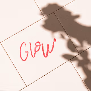 The word “Glow” written in pink lipstick across pale pink tiles with the shadow of a flower cast over part of the frame, evoking a soft, radiant mood.