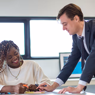Professional man in a suit assisting a smiling woman with notes during a collaborative meeting in a bright office.