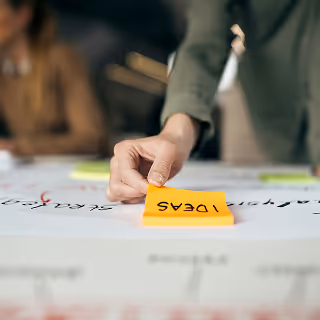 Close-up of a person's hand placing a yellow sticky note labeled "IDEAS" on a large brainstorming board during a planning session.
