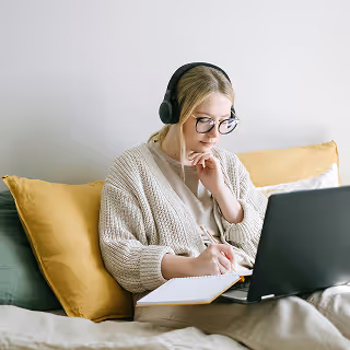 Young woman sitting on a couch with headphones on, focused on taking notes while looking at her laptop screen.