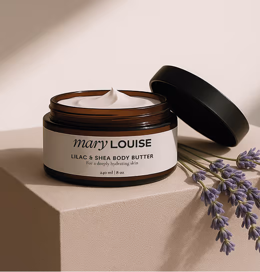 Open jar of Mary Louise Lilac & Shea Body Butter with creamy texture visible, placed on a neutral surface beside sprigs of lavender under soft natural lighting.