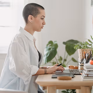 Focused woman with short hair and tattoos using a stylus at a minimalist desk, surrounded by plants, evoking a clean and modern design environment.